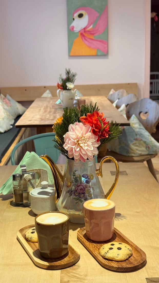 Zwei Cappuccino in Keramikbechern mit Keksen auf Holzbrettchen, dahinter eine Kanne und Blumen