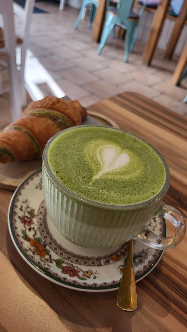 Matcha Latte mit Herz im Milchschaum, dahinter ein Matcha-Croissant auf einem Holzbrett