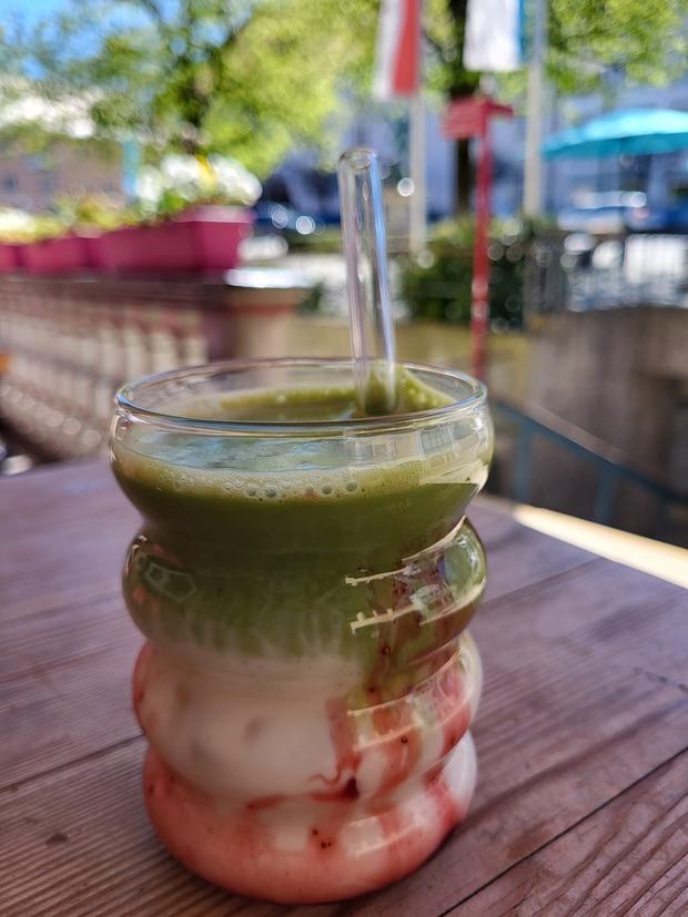 Glas mit Matcha auf Erdbeersauce und Milch, mit Glasstrohhalm, auf einem Tisch im Freien